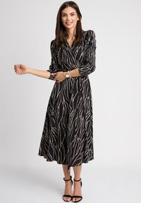 Czarna sukienka w fasonie kopertowym z białym wzorem w fale, z długimi rękawami, zaznaczoną talią i zwiewnym dołem. Połączona z czarnymi sandałami na paski.