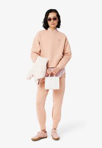 Rosa bomullstrøye med ribbestrikkede mansjetter, matchende joggebukser og rosa sneakers. Holder en liten hvit teksturert håndveske, lagdelt over en stripete skjorte.
