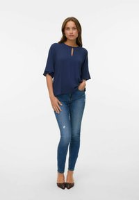 Blusa azul marino con cuello redondo y detalle en forma de ojal, combinada con unos jeans de denim azul ajustados y ligeramente desgastados, y unos zapatos marrones de punta.