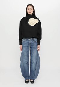 Maglione nero strutturato con collo alto e grande motivo floreale crema, abbinato a jeans a gamba larga blu e scarpe nere, su uno sfondo neutro.