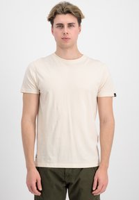 Lys beige kortermet T-skjorte laget av myk bomull, med rund hals og en liten svart logo detalj på venstre erme.