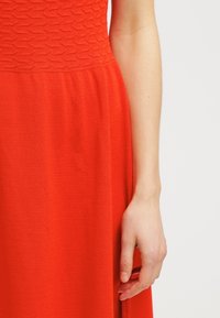 Röd klänning med en texturerad bodice med hexagonmönster, flödande kjol och ett slätt, mjukt tyg. Handen vilar på tyget.