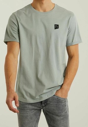 Hombre con camiseta gris claro de manga corta y pequeño logo negro en el pecho, vaqueros grises desgastados, mano en el bolsillo, fondo neutro.