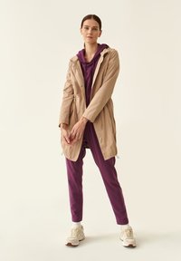 Giacca impermeabile beige con cappuccio, dotata di cordini e due tasche con zip, indossata sopra una felpa viola e pantaloni abbinati.