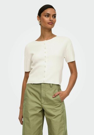 Femme portant un haut blanc à manches courtes boutonné et un pantalon ample taille haute vert olive, une main dans la poche, regardant sur le côté.