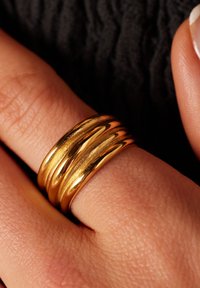 Gouden drievoudige ring met een gladde, gepolijste afwerking, met afgeronde randen en subtiele textuur, rustend op een hand tegen een donkere stof.