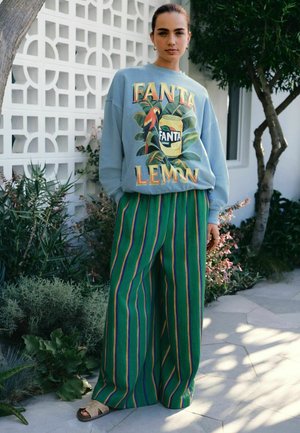 Young woman standing outdoors wearing a blue Fanta Lemon sweatshirt and green striped wide-leg pants with sandals.