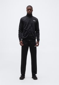 Άνδρας που στέκεται φορώντας μαύρο μπουφάν αθλητικής φόρμας Adidas, μαύρο παντελόνι φόρμας και μαύρα αθλητικά παπούτσια, σε απλό λευκό φόντο.