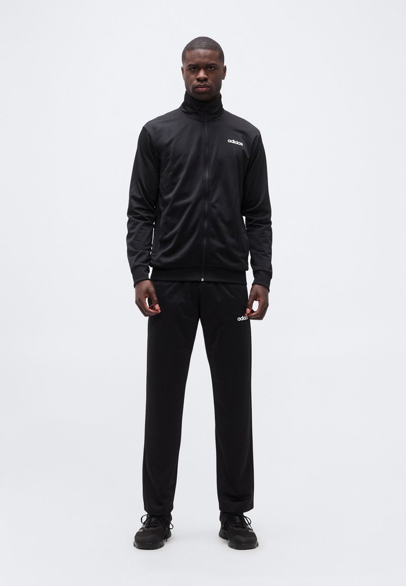 Άνδρας που στέκεται φορώντας μαύρο μπουφάν αθλητικής φόρμας Adidas, μαύρο παντελόνι φόρμας και μαύρα αθλητικά παπούτσια, σε απλό λευκό φόντο.