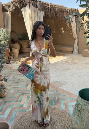 Γυναίκα με λουλουδάτο αμάνικο φόρεμα κρατάει ένα πολύχρωμο τσαντάκι και βγάζει selfie σε καθρέφτη σε μια ρουστίκ αυλή με γλάστρες και υφαντά χαλιά.