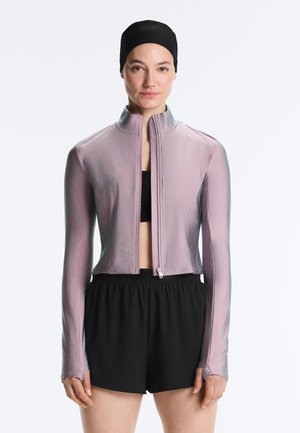 Žena v svetlofialovej veste na zips, čiernom športovom podprsenke, čiernych šortkách a čiernej plaveckej čiapke, stojaca pred jednoliatym bielym pozadím.