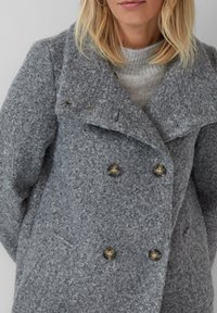 Blonde vrouw in een grijze structuur dubbele breasted wollen jas over een lichtgrijze gebreide trui.