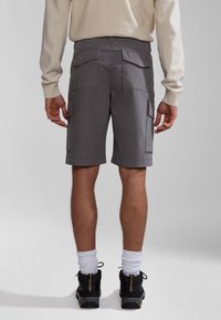 Mann, der graue Cargo-Shorts, weiße Kniestrümpfe, schwarze Wanderschuhe und ein hellbeiges Langarmoberteil trägt, vor einem einfarbigen Hintergrund steht.