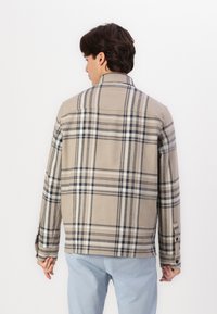 Beige plaid jacket made of soft fabric, featuring blue and white check patterns, a collar, and buttoned cuffs, worn over light blue pants.