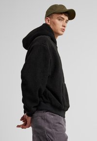 Un hoodie en polaire noire avec une grande capuche et des poches avant, associé à un pantalon en velours côtelé gris. Le modèle porte une casquette verte, se tenant devant un fond gris.