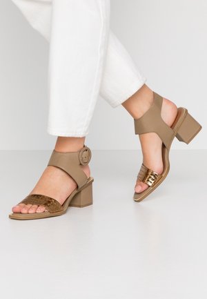 Pies con sandalias de tacón ancho beige, con tiras anchas en el tobillo y bandas metálicas texturizadas en los dedos, combinadas con pantalones blancos recortados.