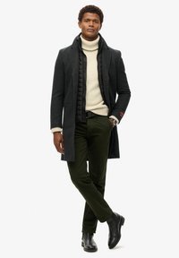 Mężczyzna stoi z założonymi rękami, ubrany w ciemnoszary płaszcz, beżowy sweter z golfem, oliwkowe spodnie, czarne buty i zegarek.