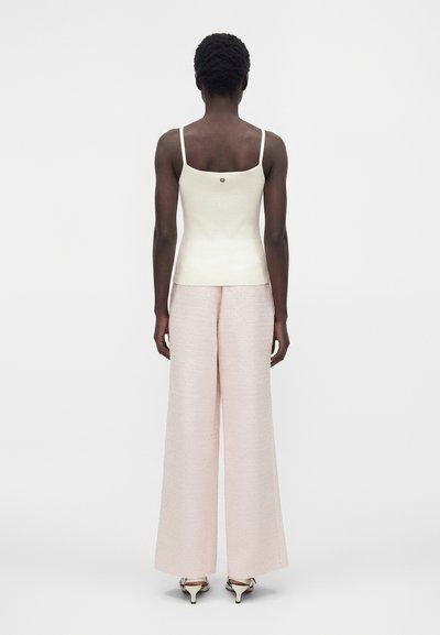 Άτομο με την πλάτη, φορούν λευκό αμάνικο τοπ, φαρδιά ανοιχτό ροζ παντελόνια και άνετα ανοιχτά παπούτσια με χαμηλό τακούνι σε απλό λευκό φόντο.