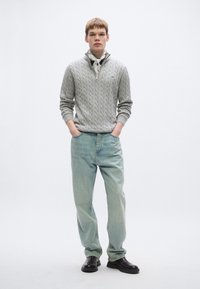 Ανοιχτό γκρι πουλόβερ με πλέξη καλωδίου και φερμουάρ, συνδυασμένο με ανοιχτό μπλε τζιν παντελόνι ίσιας γραμμής και μαύρα παπούτσια slip-on. Απλό φόντο.