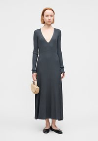 Φόρεμα με μακριά μανίκια και ριμπ ύφασμα σε σκούρο γκρι, με βαθύ V λαιμόκοψη. Φοριέται με μικρή μπεζ τσάντα χειρός και μαύρα μπαλαρίνια. Ομαλή υφή, μήκος μέχρι το πάτωμα.