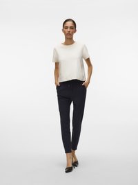 Femeie în picioare cu mâinile în buzunare, purtând un top alb cu mâneci scurte, pantaloni negri mulați și pantofi negri cu toc, pe un fundal alb.