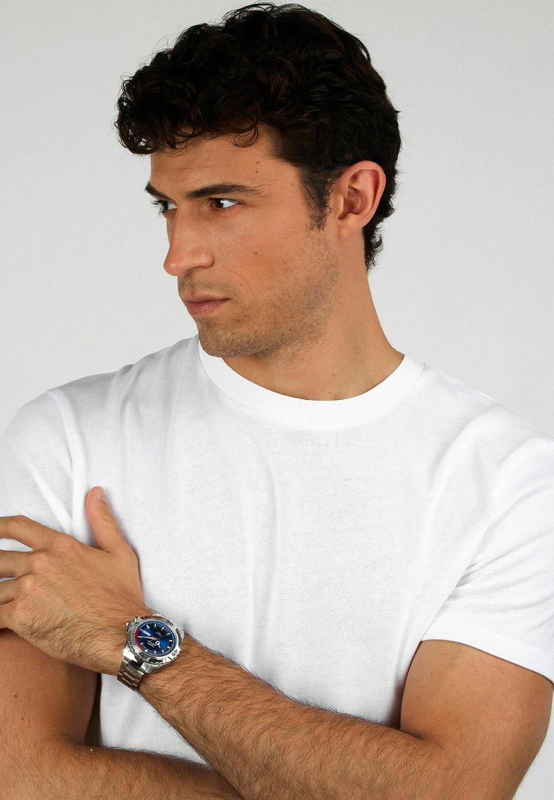 Edelstahlarmbanduhr mit blauem Ziffernblatt, silbernem Metallband und tachymetrischer Lünette. Modell trägt ein weißes Rundhalsausschnitt-T-Shirt.