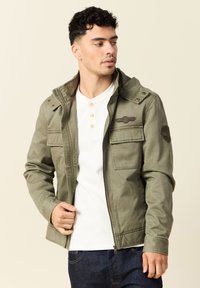 Man met een ongeopende olijfgroene jas over een witte henley-shirt en donkere spijkerbroeken, kijkend naar links tegen een effen achtergrond.