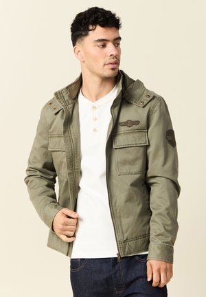 Hombre con una chaqueta verde oliva sin abotonar sobre una camiseta blanca de tipo henley y jeans oscuros, mirando hacia su izquierda frente a un fondo sencillo.