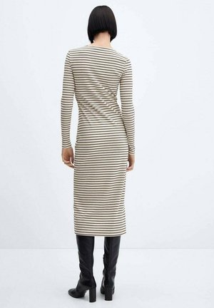 Mujer con cabello corto negro, vistiendo un vestido midi a rayas en beige y negro y botas de tacón negras hasta la rodilla, de pie contra un fondo blanco.