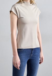 Beige, kortarmig top met een hoge hals, gemaakt van zacht materiaal. De pasvorm is iets getailleerd, gecombineerd met blauwe denim jeans.