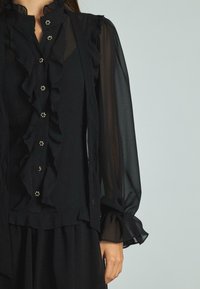 Blusa nera con parte anteriore a volant, collo alto e maniche lunghe trasparenti. Presenta bottoni decorativi con accenti floreali e polsini arricciati.