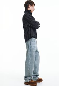 Zwart-grijs geruit overhemd, lichtblauwe loszittende jeans met witte bloemmotieven en bruine suède schoenen. Zijaanzicht van het model.