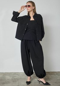 Melna oversize stila pogājama krekla ar divām krūšu kabatām, valkāta kopā ar piegulošu tvertni un vaļējām melnām biksēm. Apavi ar asu pirkstu papēžos.