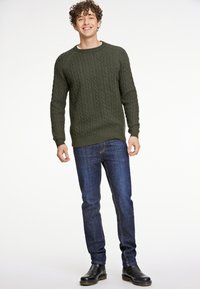 Oliwkowy sweter z dzianiny w splot kablowy, z okrągłym dekoltem, noszony na ciemnoniebieskich dżinsach i czarnych botkach. Teksturowany materiał z prążkowanymi mankietami i dołem.