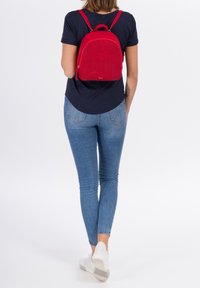 Zaino rosso con motivo floreale tagliato al laser, forma arrotondata, chiusura con zip e manico superiore. Indossato sulla schiena da una persona in camicia blu scuro e jeans.