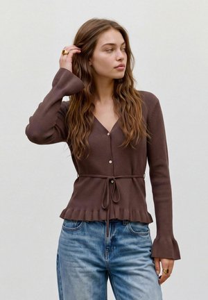 Jeune femme aux longs cheveux ondulés porte un cardigan brun boutonné avec une ceinture à la taille et un jean bleu en denim, regardant sur le côté.