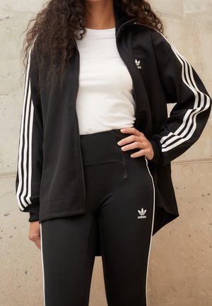 Osoba nosząca czarną kurtkę Adidas z białymi paskami i logo, białą koszulkę oraz czarne legginsy z bocznymi paskami i logo.