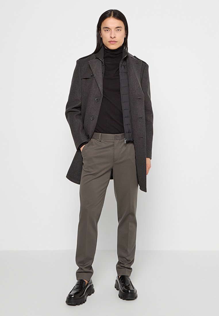 Hombre con pelo largo que lleva un abrigo gris, un suéter negro de cuello alto, pantalones oliva y mocasines negros, está de pie contra un fondo blanco.