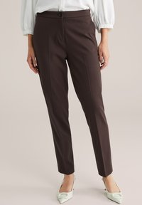 Hnedé nohavice na mieru so slim fit strihom, s jedným gombíkom na zapínanie, textúrovanou tkaninou a hladkým povrchom. Skombinované s svetlými topánkami.