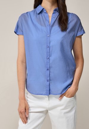 Kvinde iført en lyseblå kortærmet knap-op skjorte, der ikke er stoppet ind, over hvide bukser, stående med den ene hånd i lommen.