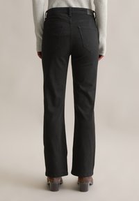 Jean flare en denim noir avec une taille mi-haute, dotés de deux poches arrière et d'une texture lisse, associés à des chaussures à talon bloc marron.