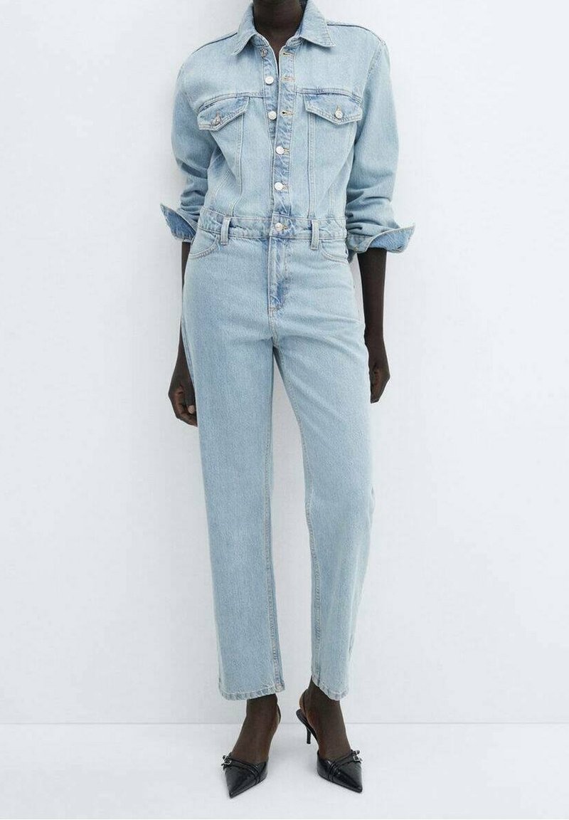 Personne portant une veste en jean bleu clair et un jean avec des manches retroussées et des escarpins noirs à bout pointu, debout devant un fond blanc uni.