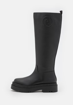 Botte haute en cuir noir avec bout rond, semelle épaisse à rainures, logo de marque embossé près du haut et languette à l’arrière.
