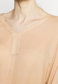 Blouse beige légère avec un décolleté en V et une texture subtile. Tissu doux qui tombe librement, présentant un petit détail en forme de clé au niveau du col.
