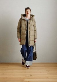 Puffer jacket in olive green, featuring a hood, zip closure, and quilted design. Paired with loose blue jeans and burgundy sneakers.