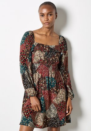 Frau, die ein langärmliges, gestepptes Kleid mit Blumen- und Tiermotiven und einem quadratischen Ausschnitt trägt und vor einer weißen Wand steht.