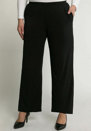 Osoba nosząca luźne czarne spodnie z bocznymi kieszeniami i czarne buty na wysokim obcasie, stojąca na jednolitym jasnym tle.