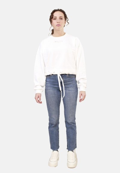 Felpa bianca corta con coulisse e logo, abbinata a jeans blu aderenti e sneaker bianche. Tessuti lisci in tutto.