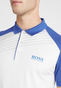 Man i vit och blå pikétröja med blå krage och "BOSS HUGO BOSS"-logotyp på vänster bröst.