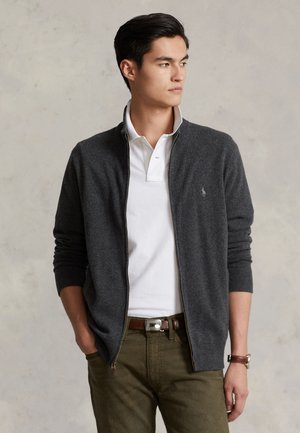 Junger Mann trägt einen grauen Zip-up-Pullover über einem weißen Poloshirt mit olivgrünen Hosen, steht mit den Händen in den Taschen und schaut zur Seite.